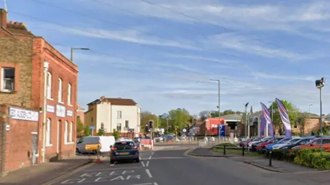 Google Part of Lower High Street in Watford. There are cars approaching a junction. On one side is a brick building and cars at a dealership parked on the right. 