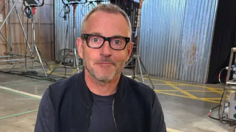 Rob Bullock sitting in the studio. He has short, brown hair and is wearing black-rimmed glasses and a black top. Corrugated iron walls and metal lights can be seen in the background.