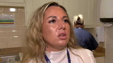 A woman with blonde hair in a kitchen talking to camera.