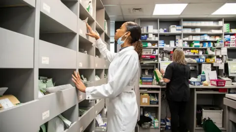 Getty Images A woman in a white lab coat in a pharmacy wearing a mask. There are numerous shelves with packs of medication on them. 