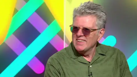 Tom Bailey has short grey hair and is sitting in a television studio wearing a loose olive-green short-sleeved shirt and lightly tinted rectangular glasses. A small microphone is clipped to the shirt. Behind the person is a brightly coloured studio backdrop featuring turquoise, yellow, pink and purple geometric shapes.