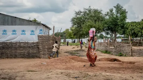 AFP via Getty Images Uma pessoa que carrega um balde rosa está andando em terreno arenoso em um campo de refugiados. Um edifício ao fundo tem chapa que diz