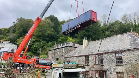 A red crane lifting a trailer that is wedged between a home and a wall. There are houses in the foreground and trees in the background.