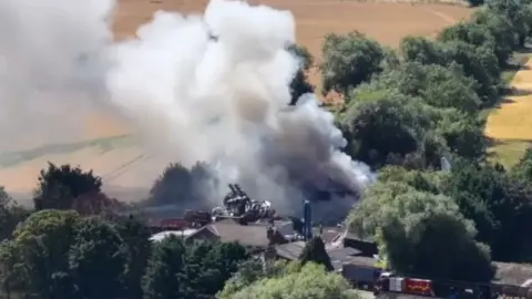 Aerial view of a large fire at a scrapyard. There is thick smoke billowing up from the blaze, and the area is surrounded by trees.