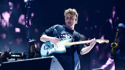 Sam Fender plays on stage with a light blue electric guitar. He has a light brown mini-mullet hairstyle and wears a black tshirt. Black and purple lighting is behind him. One man in the background, to the right, is playing a saxophone. 