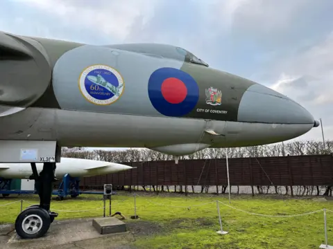 The nose of a Vulcan parked up on grass at the Midlands Air Museum. It is grey with green bands and a red circle painted below it's cockpit surrounded with a blue ring
