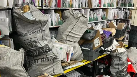A pile of Royal Mail grey sacks, full of parcels and letters, are on a shelf in front of racks of letters which ahve been sorted into bundles.