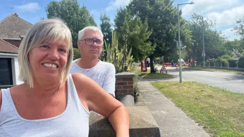 Andrew Turner/BBC Geoff and Jane May smile at the camera as they rest over their wall in front of their house. Jane has shoulder length grey and blonde hair and wears a grey vest and Geoff has short grey hair and wears a grey T-shirt.