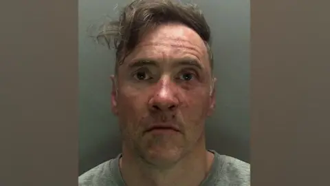 CPS A mugshot of Paul Doyle wearing a grew jumper looking stunned. His hair, a long brown fringe, is dishevelled and to the side. He is wearing a grey T-shirt.