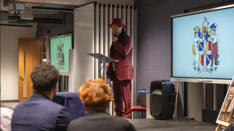 Birmingham City University A woman wearing a red suit and hat holds as a microphone as she speaks to a crowd at a podium. A large screen with the university's coat of arms is behind her.