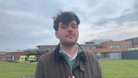 Dr Jack Fletcher has wind-swept, dark hair and is wearing a brown jacket. He is standing outside a hospital.