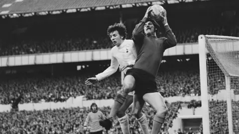 A black and white archive photo of Phil Parkes catching a ball in the air, under a challenge from Tottenham Hotspurs' Martin Peters. In the background the stands are full of fans 