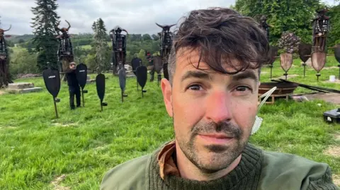 A picture of Mark Drake, who has a green jumper and has dark hair, is pictured on the Traitors set, with people working on the set behind him.