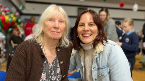 Carol and Jess look at the camera and Jess smiles. They're both sitting down and Carol is to the right. She's an older lady with a brown cardigan and a floral dress underneath. She has grey hair which is shoulder length. Jess has a denim jacket on with a cream coloured turtleneck jumper underneath. She has short dark brown hair.