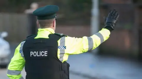 PA Media A police officer has their hand in the air. They are wearing a high visibility jacket with a black vest that has the word Police on it. They have a green hat on and a black glove. The background is blurred. 