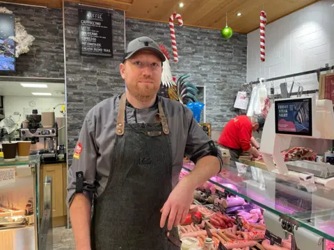 John Muff, a butcher, stands at the counter of his shop. He is wearing a grey chef's uniform and a dark apron. He is also wearing a cap. There are Christmas decorations hung above the butcher counter.