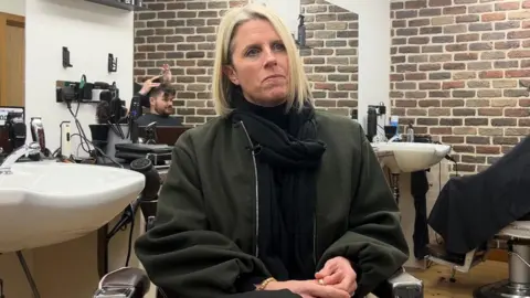 A close up shot of Lisa with long blond hair and a black jacket sat in a barber's with sinks around her and someone having their hair cut in the background