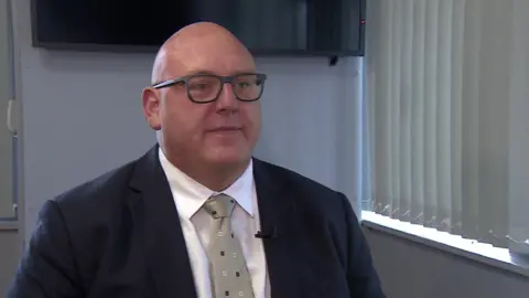 A blue suit jacket and a picture of the Vincent Hoder in a white shirt. He is wearing a brown tie with blue and white dots and glasses. 