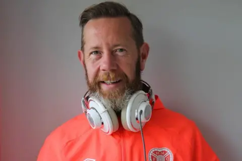 Colin Brown Colin Brown has short fair hair and smiles at the camera. He wears a fluorescent Hearts strip and has headphones around his neck