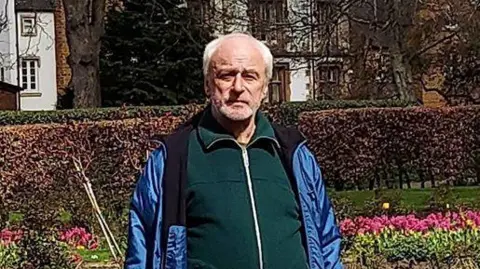 Thames Valley Police Leszek Kaminski has short grey hair and is wearing a blue coat. He is stood in a park, in front of a large flower bed.