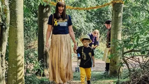 Family handout Lisa Radcliffe with shoulder length black hair, wearing a black T-shirt and a long gold pleated skirt, holding the hand of son George, aged three, who is wearing a large yellow hat, a black T-shirt and yellow trousers, while they walk through woodland