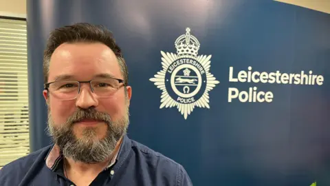A head and shoulders view of a bearded man with glasses stood in front of a banner for Leicestershire Police including the force crest.