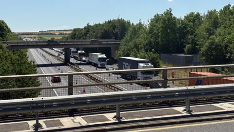 Ben Schofield/BBC View of the M11 from a bridge over the top of it. The right lanes in both directions have been blocked by orange traffic cones. The sky is blue and bright and there are green trees at either side of the motorway. The left side of the motorway is quieter than the right which has a long queue of traffic.