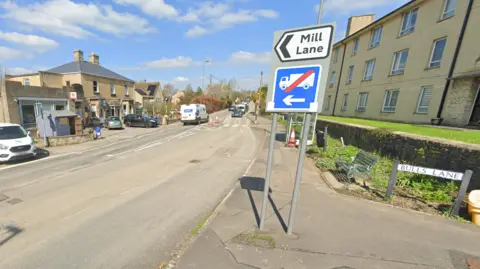 Google A signboard with two signs, one indicating Mill Lane and the other that there is no access for lorries. The signboard is next to a row of houses on the right, a Post Office outlet on the left and a zebra crossing in the distance.