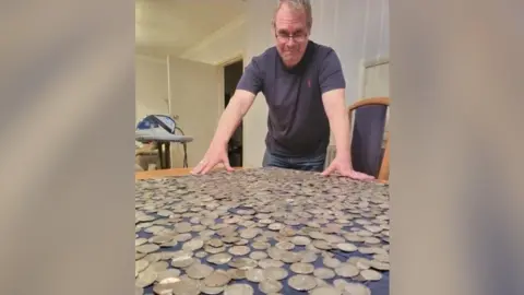 Steve Hickman Steve Hickman, a man stood over a wooden table. On the table is a blue tablecloth which has a hoard of coins on top of it. He is wearing a blue T-shirt and pair of glasses and is looking over the hoard towards the camera.