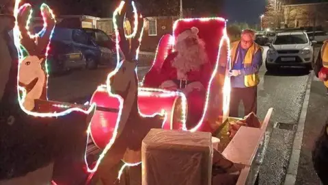 Wisbech Lions A Santa sleigh complete with two cut-out reindeers and a live Santa sitting in the driver's seat. The main red vehicle is festooned in fairy lights and a second man is at the rearmaking some adjustments to it.