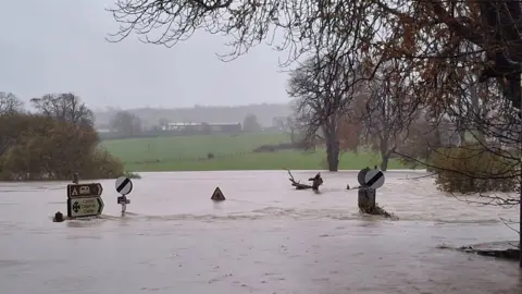 Emma Beattie Flooded Llechryd Bridge, which passes over the river Teifi in Ceredigion. Brown flood-water surges across the bridge, which is no longer visible with only the road signs appearing above the water level. 