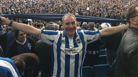 Kieran Maguire is a man with short white hair wearing a blue and white Brighton & Hove Albion football shirt. He holds a blue and white scarf over his head while in the stands at The Amex. There are thousands of people on the field.