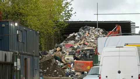 Chris Coghlan A pile of rubbish next to some trees, as well as an orange digger. There are also metal mobile site huts, a white van and a car.