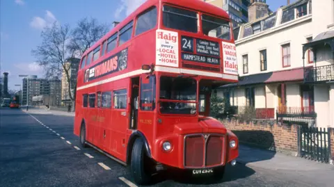 An old red Routemaster bus on a street next to houses