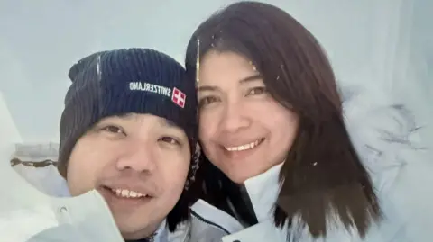 A photo of a man and woman smiling at the camera, they are both wearing white coats, the man has a navy hat on, the woman has dark hair.