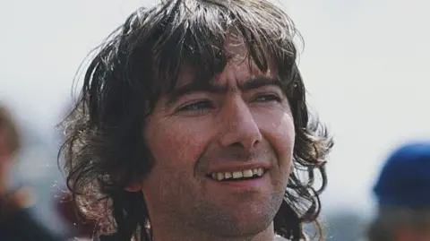 Joey Dunlop has long dark brown hair with a fringe, he has dark eyebrows and facial stubble. He is wearing a white racing suit and is looking to his left, the background is blurred.