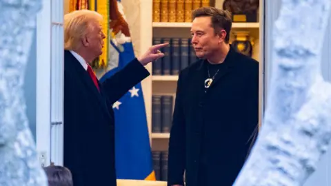 Getty Images Donald Trump and Elon Musk stand facing each other in office space as Trump gestures in conversation.