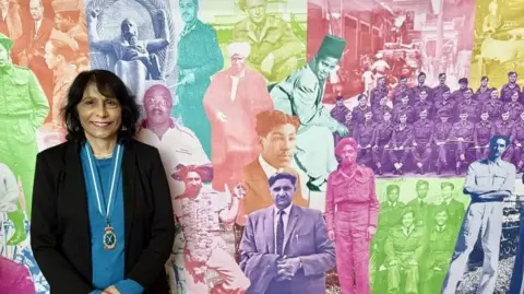 Kalpna Woolf Ms Woolf stands in front of a montage of faces making up the photo banner. Each person's picture is a different colour. Ms Woolf wears a black jacket with a blue jumper underneath. She has a medal around her neck with a blue and white tie and a gold medal.