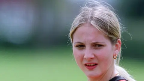 Getty Images A headshot of a blonde woman looking to the right of the camera. She has straight hair tied back in a low ponytail and gold earrings and necklace. The grass of a football pitch can be seen behind her.