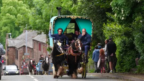 Three men ride in a carriage being pulled by two horses. It has a green top and is edged with bright fabric. Appleby's stone-built houses can seen in the background as people walk along the pavement. Other, less ornate uncovered carriages follow behind.