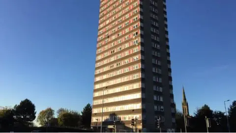 A multi-storey block of flats, which features brown stone striped with red around windows. Trees are visible around the base of the tower block and the spire of a church in the background The sky is cloudless and blue, but dark as if it is dawn or dusk. 