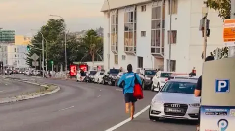 Alexandra Ching/Instagram A man wearing a blue lifeguard shirt runs barefoot down a hit towards Bondi Beach, carrying a red bag of medical supplies.