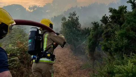 Guernsey Fire and Rescue Service Two firefighters carrying equipment and a hose. There there trees and bushes around. The sky is grey and filled with smoke.