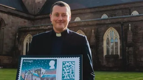 The Dean of Armagh, The Very Reverend Shane Forster, holding a small sign with the image of the stamp design on it - the stamp is an illustration of St Patrick's Cathedral with lots of children outside of it playing in the snow. The cathedral is covered in snow with the lights on and the sky has a dark sunset. The Dean is standing outside the cathedral while holding the stamp. 