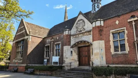 An old stone and brick building, that says Public Library at the front, with a large number of black, small paned windows. There are steps up to the entrance, with a large wooden door. There are two signs and a green tree to the left. Bushes are out the front.