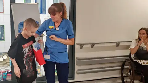 Stephanie Muirhead Andrew wearing a black t-shirt, standing next to Angela, wearing scrubs in the hospital with Stephanie looking on.