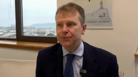 Matt Thomas sitting in an office, with a window behind him. He's dressed in a navy blue suit with a light blue shirt and bright blue tie. 