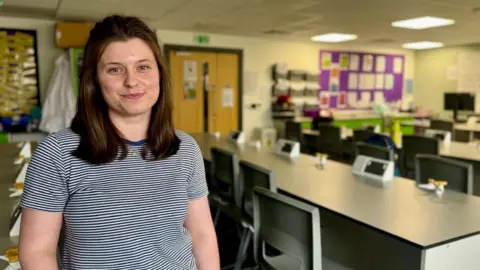 Hazel Shearing / BBC Anna Helder is seen wearing a striped T-shirt standing in a science laboratory classroom. Rows of workbenches with stools and lab taps fill the room, with noticeboards, equipment and a classroom door visible in the background.