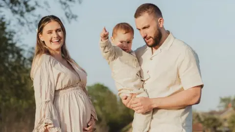Chloe and her family all wear beige and stand outside in the sunshine with trees visible in the background as part of a professional family photoshoot. Chloe is visibly pregnant and holds her stomach smiling, while her partner holds their son.