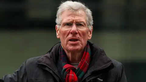 Getty Images Kenny MacAskill has short grey hair and wears silver wire rimmed glasses. He wears a red and green tartan scarf and black rain jacket and looks directly into the camera